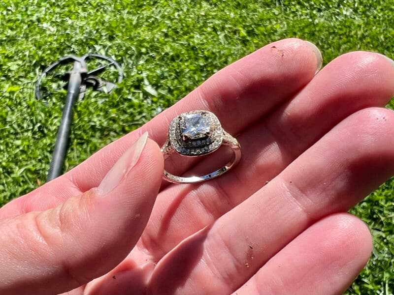 A diamond ring held between two fingers, with a metal detector on the grass behind it.